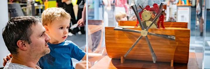 Bild eines Vaters mit seinem circa vierjährigen Sohn die auf ein in einer Vitrine liegendes Modell aus der Da Vinci Ausstellung schauen. Das Modell zeigt einen Schaufelradantrieb anhand eines Zahnradgetriebes und einem aufgeschnittenen Bootsrumpf