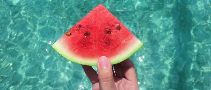 Hand hält ein Stück Wassermelone über Poolwasser