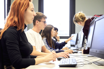 Vier Studierende sitzen in einem Computerraum an Computern und eine Person lehnt sich zu einer Studierenden runter um ihr zu helfen