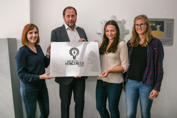 drei Studentinnen mit dem Dekan in der Mitte halten ein silbernes Schild in die Kamera