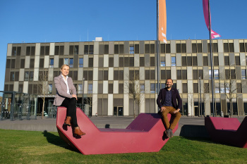 Eine Frau und ein Mann sitzen bei Sonnenschein auf einer Sitzgelegenheit vor dem FH-Hauptgebäude in Bielefeld.