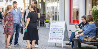 Eine im Kreis stehende Gruppe Menschen steht vor der zur Science Bench umfunktionierten Bank und unterhält sich Auf der Bank sitzen Heike Wulf und Karin Hamel und sind ins Gespräch vertieft