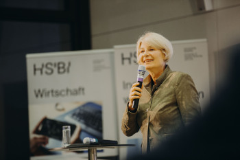 Die Präsidentin der HSBI Professorin Dr Ingeborg Schramm Wölk steht in einem olivgrünen Blazer vor einem Banner mit dem Aufdruck des Fachbereichs Wirtschaft und hält ihr Grußwort an die Besucherinnen des Demo Days