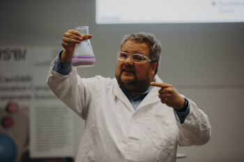 Bild von Matthias Pieper während seines Pitches Er steht in einem weißen Laborkittel und mit Schutzbrille auf der Bühne des Demo Days In seiner rechten Hand hält er einen Erlenmeyerkolben in dem sich eine lila Flüssigkeit mit einer weiteren Phase befindet