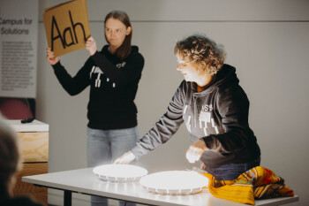 Katharina Schnatmann steht links im Bild und hält in einem schwarzen Kapuzepullover ein Schild hoch auf dem Aaah steht Heike Wulf steht rechts von ihr auf dem Tisch vor ihnen liegen eingeschaltete Lampen