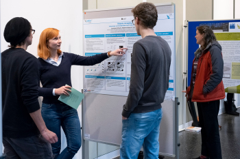 Eine Frau steht an einem Poster und erklärt anderen Personen etwas.