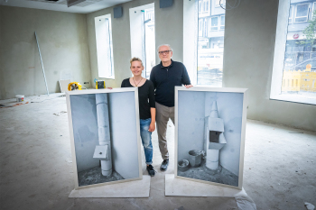 Doppelporträt von Kim Groche und Professor Roman Bezjak vom Fachbereich Gestaltung auf der Baustelle des Ausstellungsraums Satellit Vor ihnen stehen großformatige gerahmte Bilder als Anschuungsobjekte