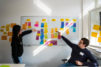 Zwei Personen stehen vor einer weißen Tafel mit bunten Post its