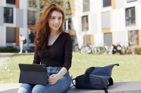 Eine Frau sitzt mit einem Tablet auf einer Steinbank, neben ihr ein Rucksack