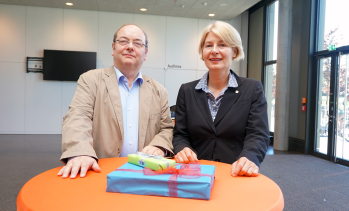 Prof. Dr.-Ing. Ralf Hörstmeier und FH-Präsidentin Prof. Dr. Ingeborg Schramm-Wölk. stehen an einem Stehtisch, auf dem ein Geschenk liegt.