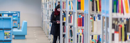 Studierende steht vor einem Regal in der Bibliothek und liest in einem Buch