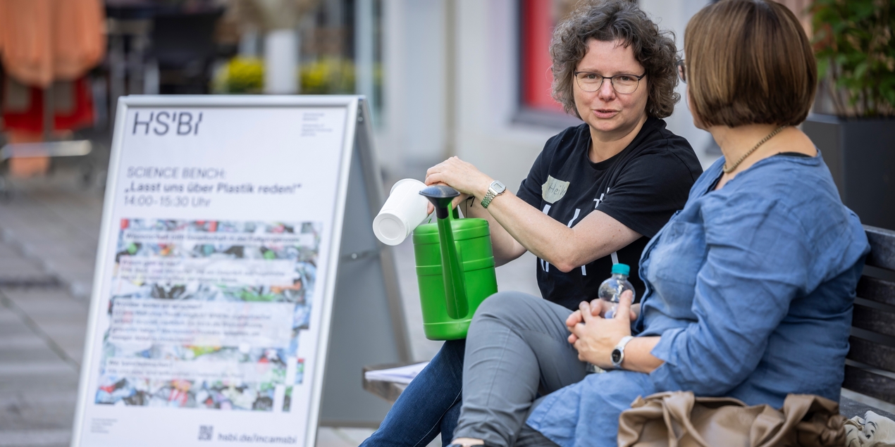 Im Vordergrund sieht man einen Plakataufsteller mit einem Plakat der Science Bench Aktion dahinter sitzt Heike Wulf aus dem Incams@BI Projekt und unterhält sich mit einer rotblonden Frau