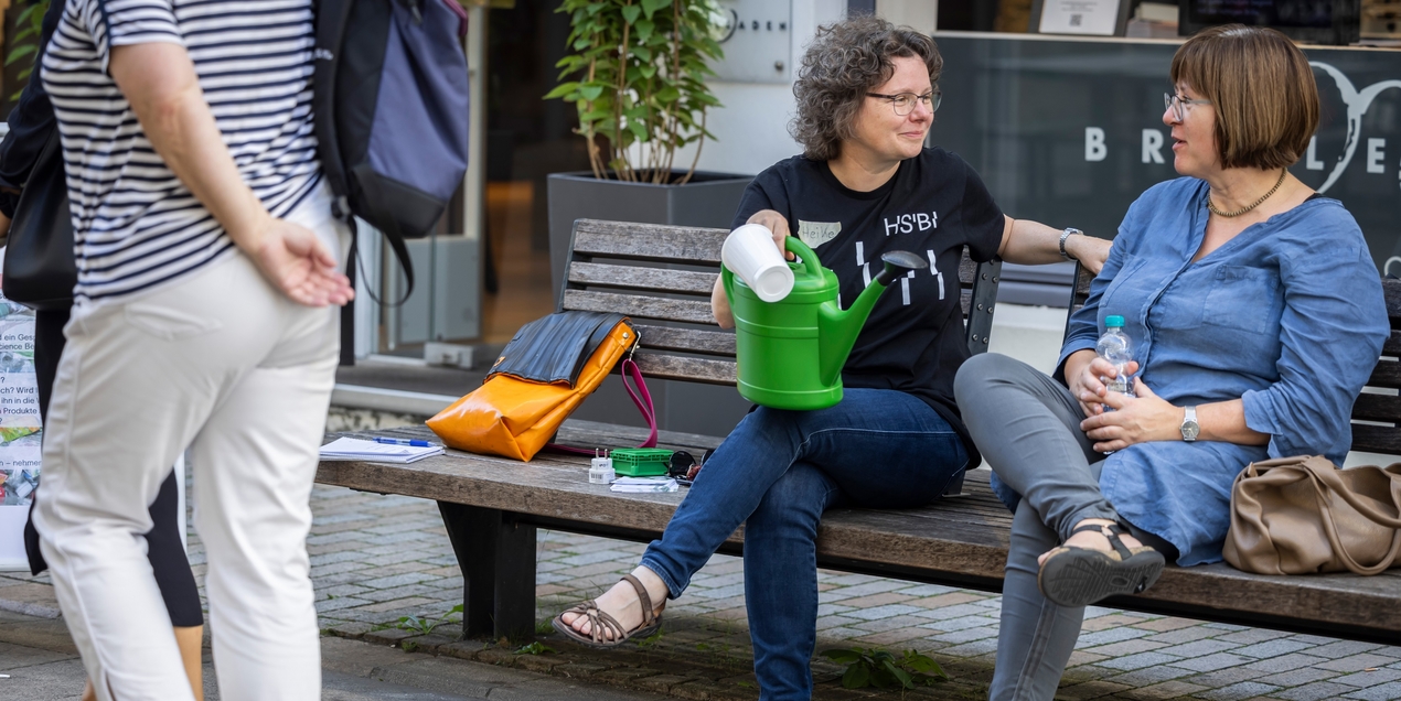 Heike Wulf sitzt in einem schwarzen T Shirt auf einer Bank und spricht mit Karin Hamel Beide haben ihre Beine übereinandergeschlagen und lächeln sich an