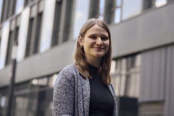 Porträtbild von Katrin Handel die in einer grauen Strickjacke vor dem Gebäude der HSBI steht Sie hat hellbraune Haare und lächelt im Halbprofil in die Kamera