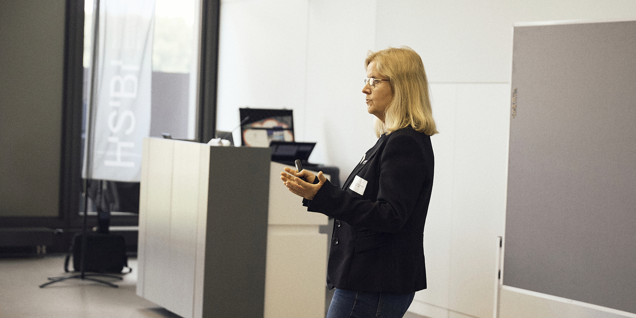 Bild von Professorin Doktor Eva Schwenzfeier-Hellkamp die in einem schwarzen Blazer vor einer grauen Pinnwand im Konferenzsaal der HSBI steht und das Projekt InCamS@BI vorstellt