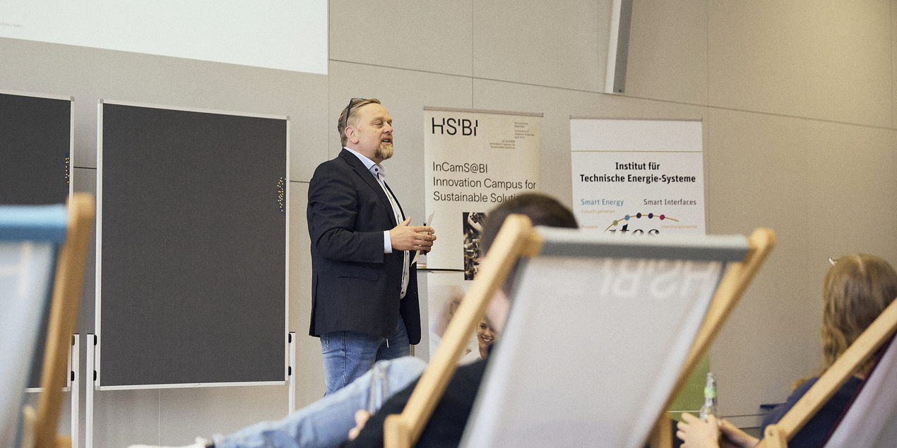Bild von Professor Jens Haubrock der in einem dunkelblauen Jackett im Konferenzbereich der HSBI steht Er hat seine rechte Hand leicht angehoben Hinter ihm steht ein Student in beigem T Shirt und lacht