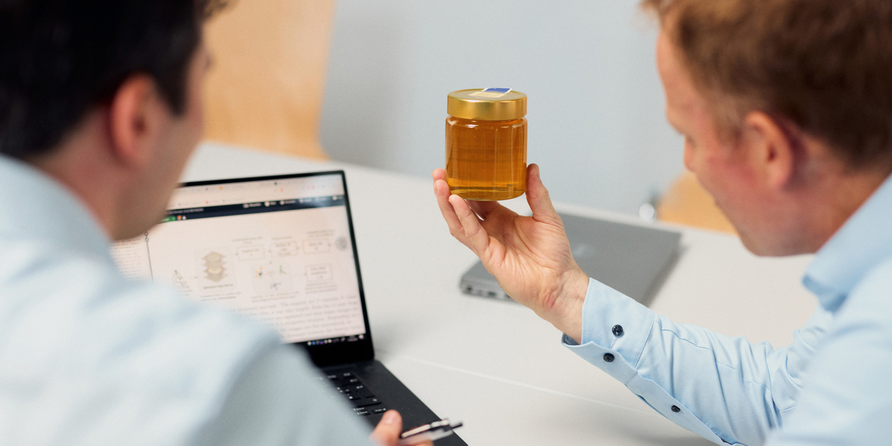 Zwei Personen blicken auf ein Glas Honig, dahinter steht ein Laptop