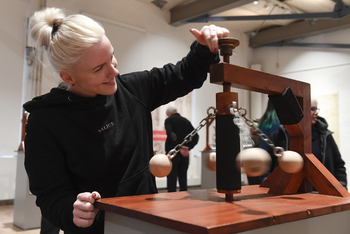 Eine Frau steht vor einer Konstruktion aus Holz mit Kugeln an Ketten auf einem Tisch