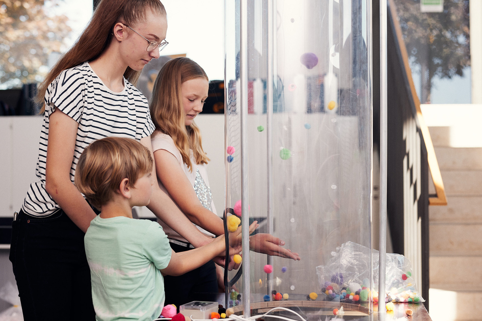 Drei Schüler*innen lassen bunte Kugeln in einem Windkanal schweben.