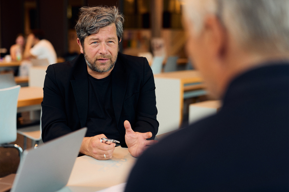 Professor Oliver Bierhoff sitzt in einem schwarzen Sakko und einem schwarzen T Shirt an einem Tisch in der Cafeterie der HSBI Während er spricht blickt er eine ihm gegenübersitzende Person an