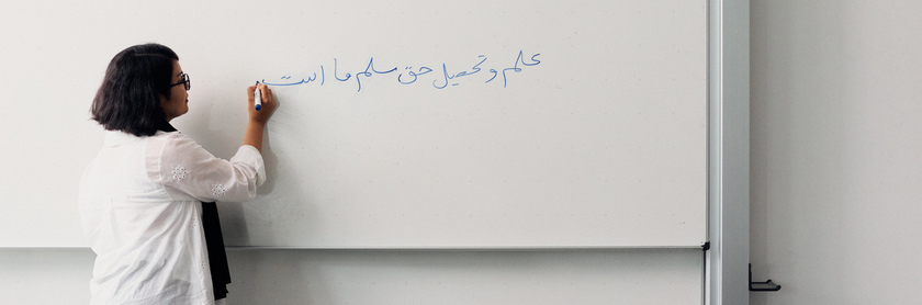 Farkunda Karimi steht mit dem Rücken zur Kamera Sie schreibt mit einem blauen Filzstift an eine weiße Tafel in arabischer Schrift Auf der Tafel steht der Satz Wissenschaft und Bildung sind unser unveräußerliches Recht