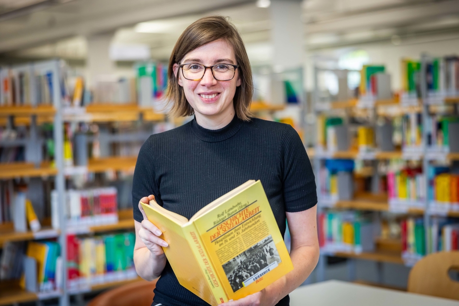 Sandra Holtgreve steht in der Bibliothek und hält ein Buch in die Kamera