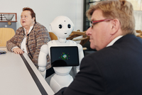 Zwei Personen sitzen an einem Tisch, zwischen ihnen der Roboter Pepper.