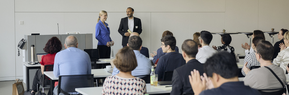 Natalie Bartholomäus and Riza Öztürk stand next to the lectern and speak to the participants