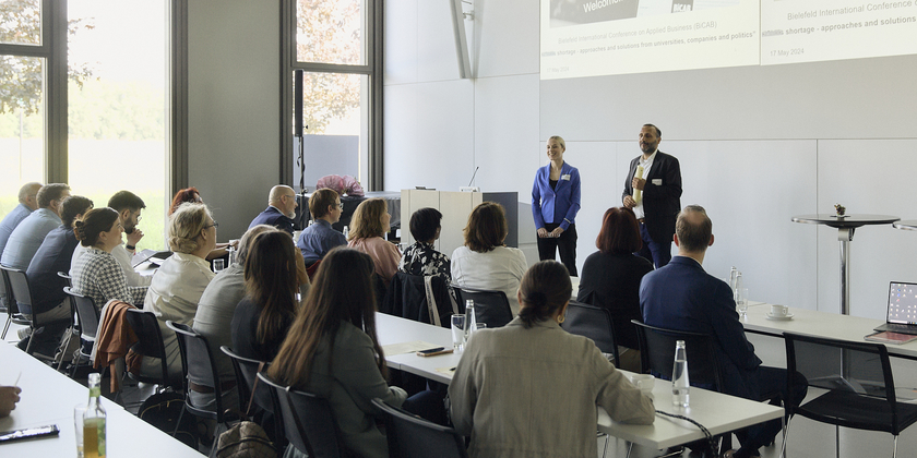 Natalie Bartholomäus and Riza Öztürk stand next to the lectern in the conference hall and address the participants