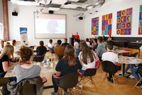 Eine Gruppe von Personen sitzt an Tischen und blickt nach vorne zu einer Präsentation.