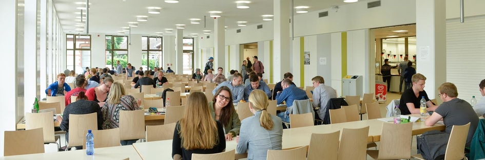 In einem großen Saal mit vielen Tischreihen sitzen Studierende, die essen und sich unterhalten.